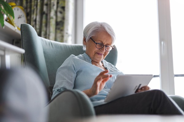 Elderly woman sits in a big chair using a tablet connected to the internet.