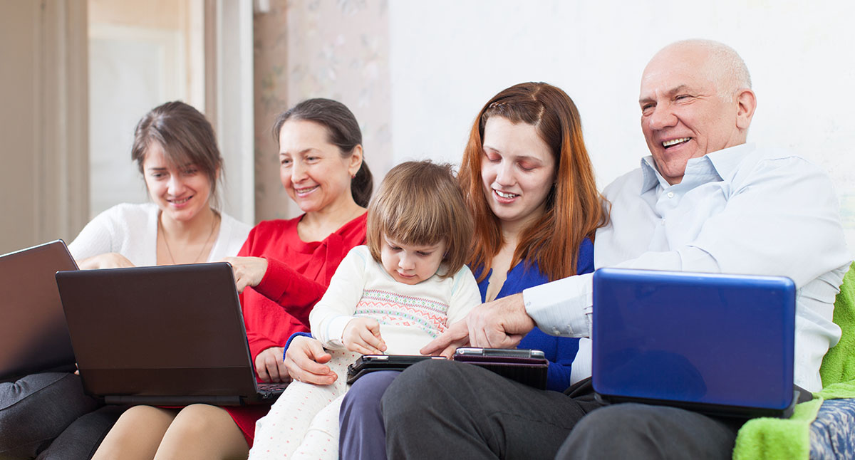 A family with diverse ages sits on a couch each holding a laptop while a child holds a tablet.