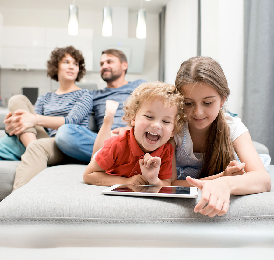 Happy children play on a tablet with their parents smiling in the backgroound.