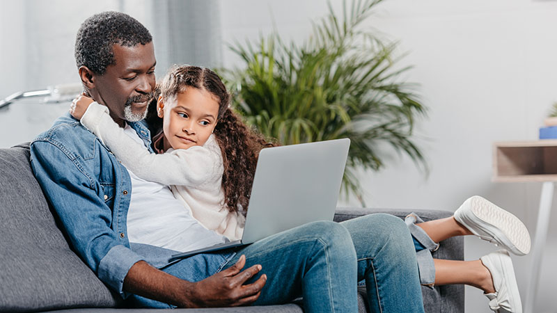 A father and daughter sit on the couch using a laptop.