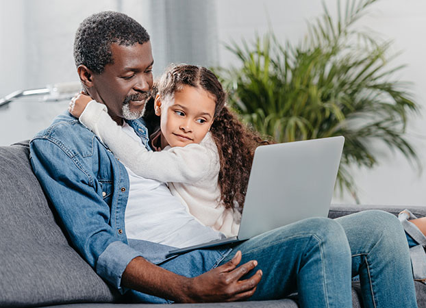 A father and daughter sit on the couch using a laptop.