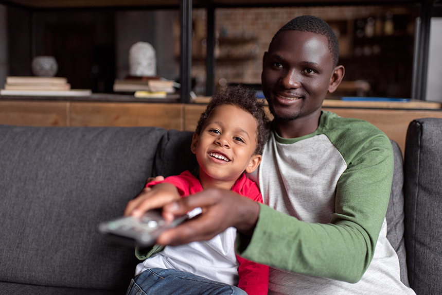 A father and son sit on a couch each with a hand on a television remote while smiling.