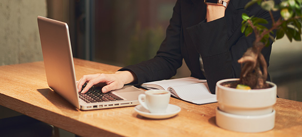 A cropped images of a woman typing on her laptop holding a coffee.