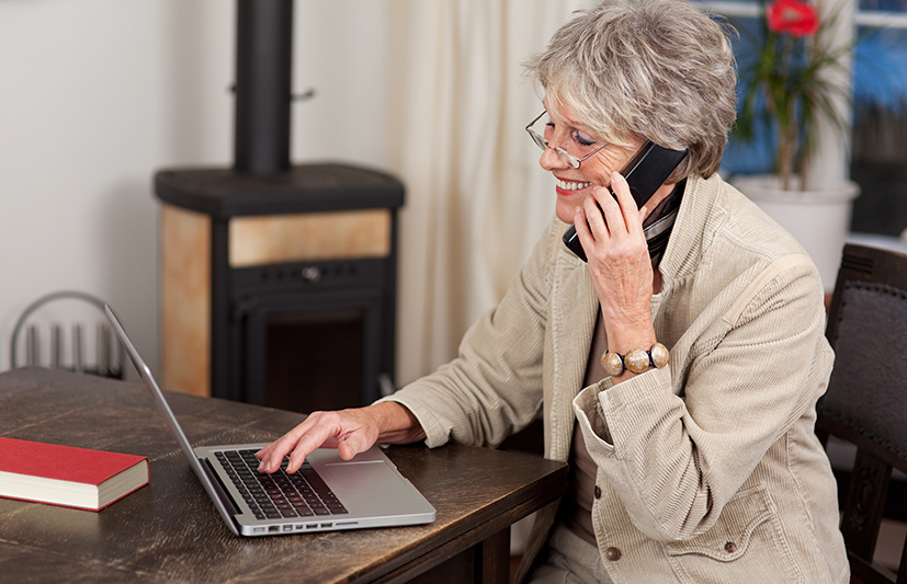 An elderly woman smiles while talking on the home phone and using a laptop.